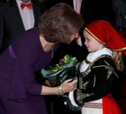 Doña Sofía recibe un ramo de flores de manos de una niña ataviada con un traje típico griego